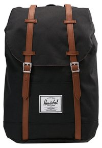 Sac à dos en toile noire avec des sangles et des accents en cuir marron, doté d'une poche avant avec une étiquette de logo blanc et une fermeture à rabat.