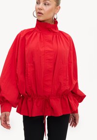 Femme portant une veste rouge ample avec un col montant, une taille froncée et de longues manches bouffantes, associée à un pantalon noir et des boucles d'oreilles créoles.
