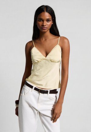 Donna con lunghi capelli neri che indossa una canottiera giallo pallido e pantaloni bianchi con una cintura nera, in piedi davanti a uno sfondo grigio uniforme.