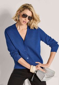 Vrouw met kort blond haar in een zonnebril en een blauwe textuur cardigan, die een zilveren handtas vasthoudt, staande tegen een lichte achtergrond.