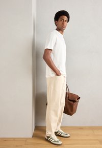 White short-sleeve t-shirt, beige relaxed-fit pants, and green-striped sneakers. Brown leather bag with top handle. Wooden floor backdrop.