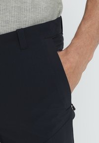 Pantaloni blu navy realizzati in tessuto leggero, dotati di tasca laterale, passanti per la cintura e una texture liscia. Camicia grigia a motivo parzialmente visibile.