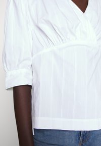 Hvit, stripsbluse med korte, puffete ermer og samlet midje. Stoffet ser glatt ut med en lett glans. Kanten er rettskåret.
