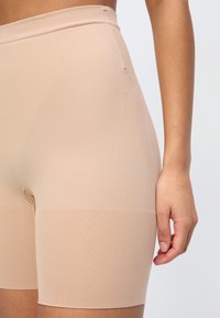 Μπεζ αδιάκοπο σορτς σχήματος με ψηλή μέση, φορεμένο στο κάτω μέρος του κορμού, που εκτείνεται μέχρι τα μέσα μηρών, εμφανίζεται σε απλό ανοιχτό φόντ ο.