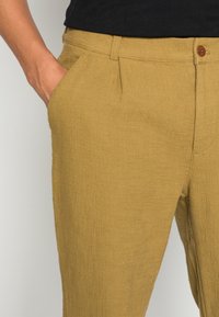 Pantaloni gialli a trama con tasche laterali, chiusura con bottone e una vestibilità leggermente strutturata, mostrati con un top nero parzialmente visibile.