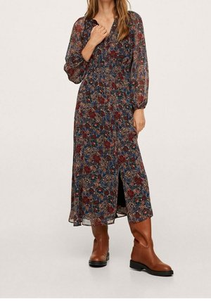 Femme portant une longue robe à motif floral avec des manches longues en voile et des bottes en cuir marron jusqu'au milieu du mollet, debout devant un fond uni.