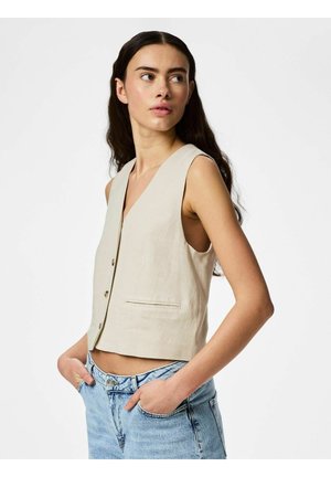 Noor naine pikkade tumedate juustega, kandes varrukateta beežikat vest ja helesiniseid pükse, käed taskutes, vaatab kõrvale.