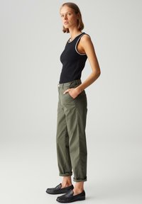 Top senza maniche nero con rifiniture bianche, abbinato a pantaloni verde oliva a vestibilità rilassata e scarpe nere slip-on, che mostrano una texture dettagliata.