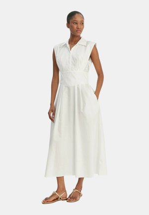 Donna che indossa un vestito midi bianco senza maniche con colletto e tasche laterali, abbinato a sandali piatti beige, in piedi su sfondo bianco.