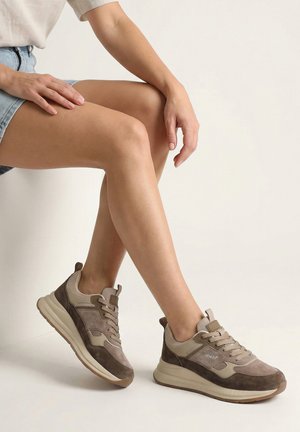 Žena v béžových a hnědých teniskách s lehkými džínovými šortkami, sedící s překříženýma nohama na jednobarevném světlém pozadí.