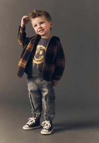 Laps, kellel on seljas pruun ruuduline jope halli graafilise t-särgi, hallide cargo-pükside ja mustade kõrgete tennisekingade valgete paeltega.