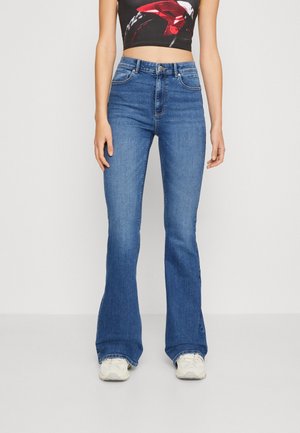 Femme portant un jean évasé bleu taille haute, des baskets blanches et un crop top noir avec un motif graphique rouge, debout sur un fond blanc uni.