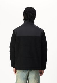 Black fleece jacket with a smooth upper panel, high collar, textured fabric, and a relaxed fit. Paired with light blue distressed jeans.