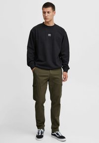Czarna bluza z okrągłym dekoltem i detalami z metką, zestawiona z oliwkowymi spodniami cargo z bocznymi kieszeniami i zwężanym krojem. Czarne sneakersy.