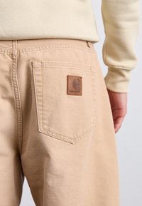Pantalones beige con un bolsillo trasero que cuenta con una parche de cuero marrón. Textura suave y ajuste estándar. Tela ligera y duradera.