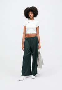 Balta sporta blūze ar īsām piedurknēm un melnu logotipu, tumši zaļas platās bikses. Modelis tur gaiši pelēku somu. Balti apavi redzami.