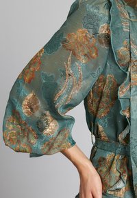 Tela transparente en tono teal con bordados florales en tonos dorados y naranjas, con mangas voluminosas y acentos con volantes.