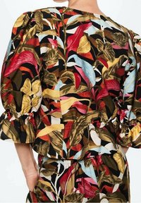 Donna che indossa un completo di blusa e pantaloni con maniche voluminose e una stampa di foglie tropicali colorate nei toni del rosso, giallo, blu, marrone e nero.