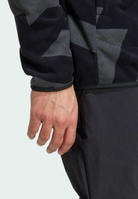 Fleecejacke mit einem mehrfarbigen, dunkelgrauen und schwarzen geometrischen Muster, ausgestattet mit einem Reißverschlusskragen sowie elastischen Bündchen und Saum.