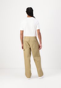 Beige cargobyxor med lös passform, två stora bakfickor och sidofickor, i kombination med en vit t-shirt med rund halsringning, sedd bakifrån.