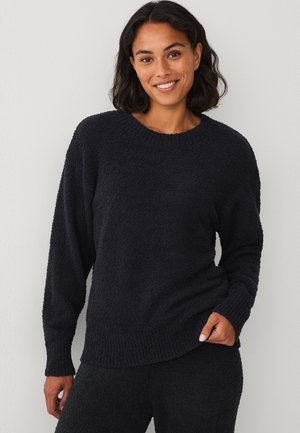 Kvinde med mørkt, mellemlangt hår iført en tekstureret sort langærmet sweater og matchende sorte bukser, der smiler til kameraet.