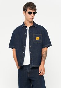 Temno denim s kratkimi rokavi s prsno vrečko, oranžna oznaka in bele gumbe. Obučeno z enobarvno belo srajco spodaj in temnimi sončnimi očali.