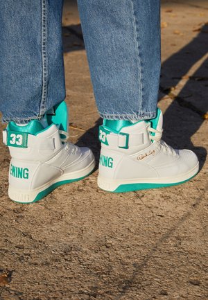 Białe i turkusowe trampki za kostkę z napisem „33” i „EWING”, noszone z niebieskimi dżinsami na teksturowanym, zewnętrznym podłożu.