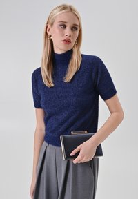 Donna con capelli biondi lisci indossa un maglione blu navy a maniche corte, pantaloni grigi a pieghe, orecchini a cerchio dorati e tiene in mano una borsa a clutch nera.