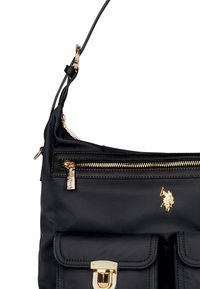 Sort nylon håndtaske med guld hardware detaljer, udstyret med en top lynlås, frontlomme med klip og et smalt justerbart rem.
