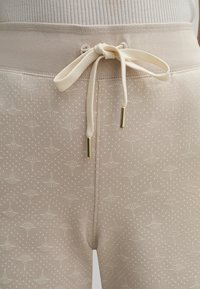 Pantalon de survêtement couleur crème avec un motif texturé de petits éventails et points, doté d'une taille côtelée et d'un cordon de serrage avec des embouts dorés.