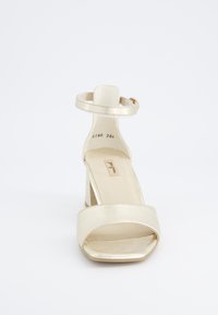 Zlaté sandály na vysokém podpatku se čtveratou špičkou, páskem kolem kotníku a blokovým podpatkem. Hladký, lesklý materiál s minimálním prošíváním.