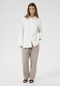 Gestreifte langärmelige Bluse in Weiß und Hellgrün, mit einer Tasche, kombiniert mit lockeren beigen Hosen und schwarzen Sandalen.