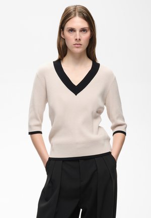 Ung kvinde med lige lysebrunt hår har en beige V-hals sweater med sort kant og sorte plisserede bukser, hænderne i lommerne.