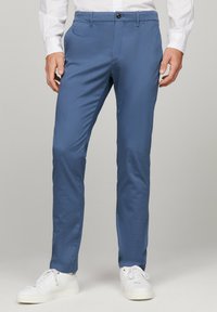 Pantaloni in cotone slim-fit blu con una texture liscia e frontale piatto. Presentano una chiusura a bottone e tasche laterali. Indossati con sneakers bianche.