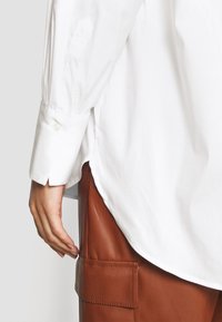 Main portant une chemise blanche à manches longues avec poignet boutonné, reposant près de la poche d'un pantalon en cuir marron avec des coutures visibles.