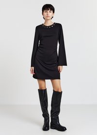 Schwarzes, langärmliges Kleid mit rundem Ausschnitt und kreisförmigen Akzenten. Glatter Stoff, figurbetontes Design, kombiniert mit kniehohen schwarzen Stiefeln.