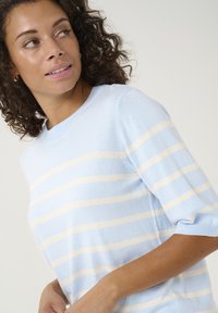 Heled sinine triibuline sviiter, millel on kreemjad horisontaalsed triibud, ümmargune kaelus ja lühikesed varrukad. Valmistatud pehmest, tekstureeritud kangast.