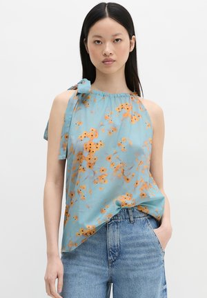 Giovane donna che indossa un top floreale azzurro chiaro senza maniche con fiori arancioni e jeans blu, in piedi contro uno sfondo bianco semplice.