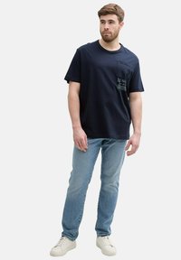 Granatowa bawełniana koszulka z kieszonką na piersi i graficznymi akcentami. Połączona z jasnoniebieskimi dżinsami i białymi trampkami.