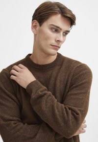 Brauner Strickpullover mit rundem Ausschnitt und gerippten Ärmelbündchen. Der Stoff wirkt weich mit einer strukturierten Oberflächenbeschaffenheit. Das Modell posiert mit verschränkten Armen.