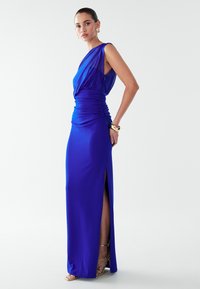 Zila vakarkleita ar vienu plecu, pieguļošu tekstūru viduklī un sānu šķēlumu. Gluda auduma un grīdas garuma siluets.