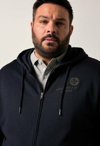 Navy zip-up hoodie with a gray shirt underneath, featuring an embroidered compass design and text "ADVENTURE IS MY COMPASS." Soft material.