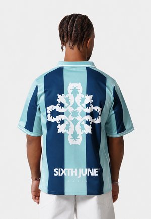 Man met ingevlochten haar draagt een blauw gestreept T-shirt met korte mouwen met een wit sierlijk ontwerp en de tekst "Sixth June Paris" op de rug.