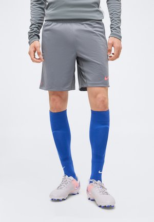 Personne portant un short de sport gris, des chaussettes bleues montantes jusqu'aux genoux, et des chaussures de soccer de couleur claire avec des accents roses et violets.