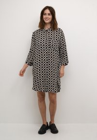 Loose-fitting cream dress with a black diamond pattern, three-quarter sleeves, button front, and asymmetrical hem. Paired with black shoes.