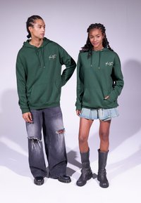Sweatshirt à capuche verte avec poche avant, porté avec un jean gris déchiré et une jupe en denim. Le modèle porte des bottines noires. Design simple.
