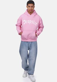 Rosa Kapuzenpullover mit frontaler Tasche und weißem "Dropsie"-Text, kombiniert mit hellblauen, locker sitzenden Jeans und weißen Sneakers.