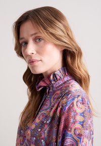 Seidenhemd mit hohem Rüschkragen und einem lebhaften Paisleymuster in Lila-, Rosa- und Goldtönen. Weiche, glatte Textur.