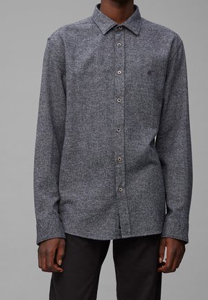 Mężczyzna w długim rękawie, szarej teksturowanej koszuli na guziki i czarnych spodniach na jednolitym szarym tle.