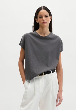 Femme aux longs cheveux noirs portant un haut gris ample à manches courtes, rentré dans un pantalon blanc taille haute avec une ceinture noire, debout devant un fond uni.